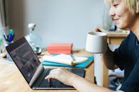 一位开朗的亚洲女性侧坐在桌旁，一边喝着热饮，一边使用笔记本电脑