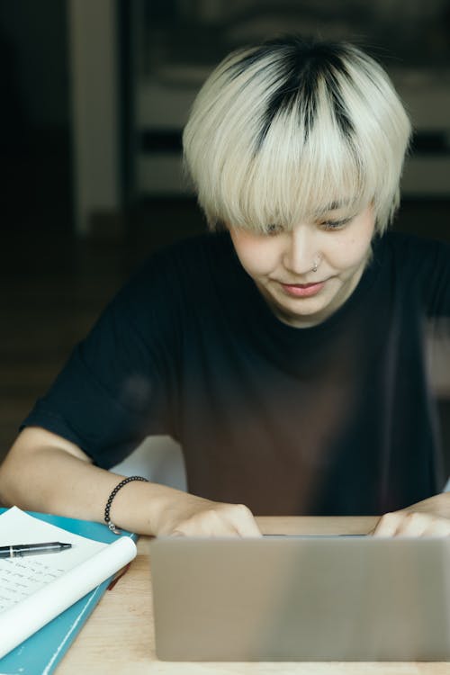 Young concentrated Asian female in black tee shirt using laptop while sitting at table with notes