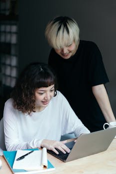 Young happy Asian girlfriends in home wear browsing laptop while gathering at wooden table in daylight