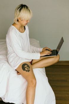 一名神情严肃、身穿白色休闲服、右臀部有纹身的女性，白天坐在舒适的床上，正浏览着上网本的侧面图
