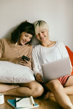 几位乐观微笑的亚洲年轻姐妹坐在地板上，戴着耳机听音乐，手里拿着笔记本、智能手机和上网本浏览信息