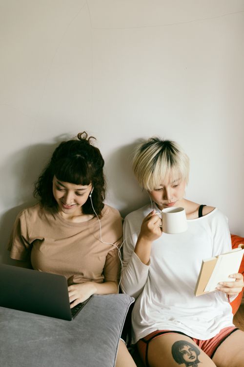 Free Content young Asian sisters in casual wear sharing earphones and sitting on floor with cup of hot drink while working on laptop and reading book together Stock Photo