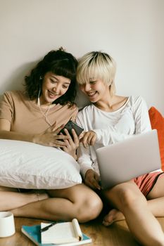 一位穿着休闲装的年轻亚洲女友坐在地板上，戴着耳机操作笔记本电脑，同时在家用智能手机浏览内容，脸上洋溢着快乐的笑容