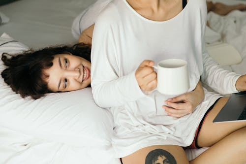 Free Cheerful young Asian lesbian couple in sleepwear resting on bed and hugging while and browsing netbook and having cup of hot drink Stock Photo