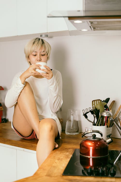 Free Young Asian female sitting on counter top with mug of hot drink in morning time in kitchen Stock Photo