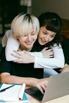 一位面带微笑的年轻亚洲女性坐在桌旁使用笔记本电脑，她快乐的朋友从身后拥抱了她