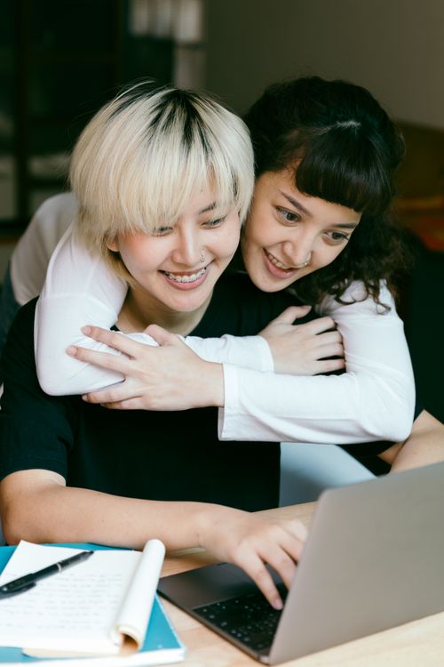 Free Young smiling Asian woman sitting at table and using laptop while happy female friend hugging from behind Stock Photo