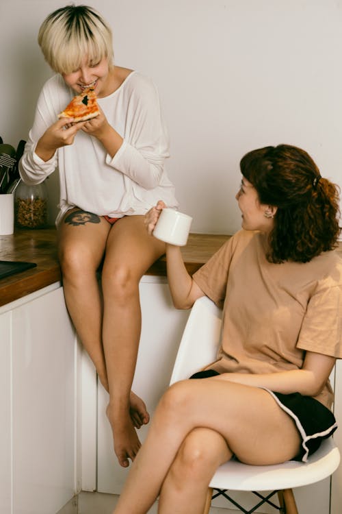 Happy tattooed blond woman eating pizza while looking at brown haired female friend with cup of coffee sitting with crossed legs during conversation in flat