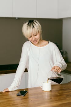 Cheerful Asian lady in casual wear and earbuds listening to music from smartphone while pouring freshly brewed coffee into cup on wooden table in kitchen at home