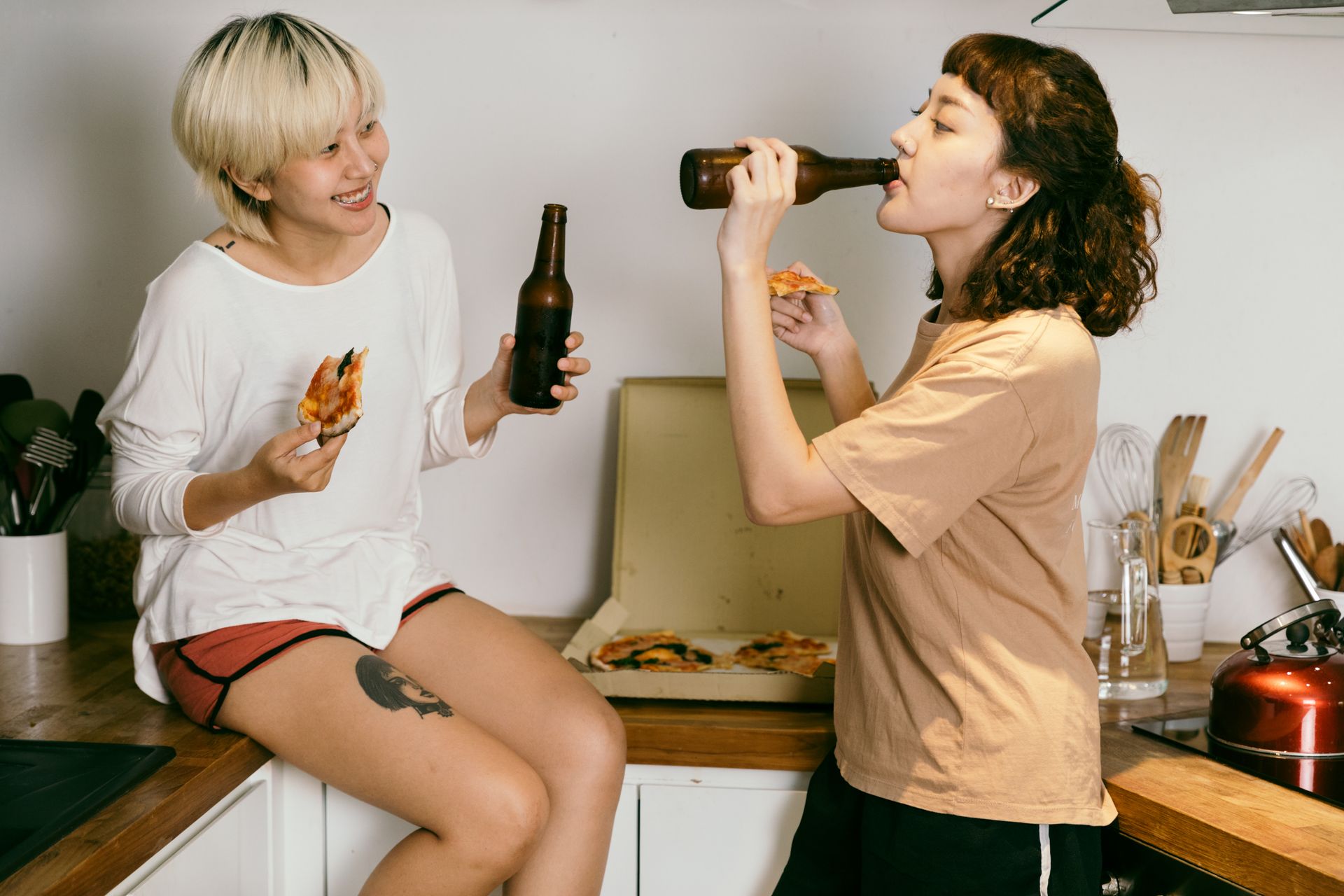 Happy tattooed blond lady eating pizza while looking at female friend in casual wear drinking beverage from bottle near kitchen utensils in apartment