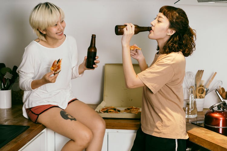 Girlfriends Eating Pizza And Drinking Beer In Kitchen At Home