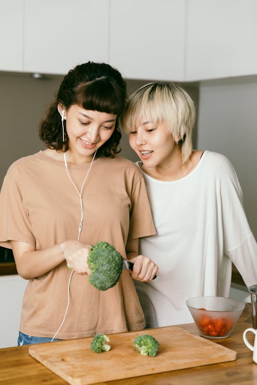 Cheerful young lady in earbuds cutting fresh broccoli for salad while standing near positive blond girlfriend in casual wear in kitchen at home