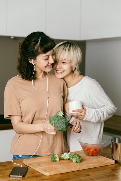Free Cheerful woman in earphones listening to music from smartphone and cutting broccoli while blond girlfriend drinking coffee in kitchen in apartment Stock Photo