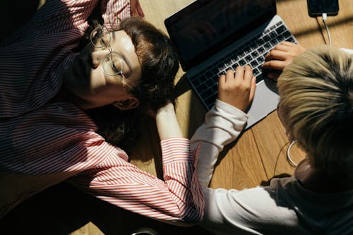 From above of lady in eyewear lying on wooden floor near anonymous female remote worker typing on netbook and listening to music in earphones form smartphone