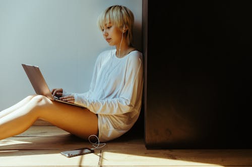 Side view of young female remote employee in casual wear and earphones listening to music while surfing internet on netbook and sitting on floor leaned on wall in sunlight