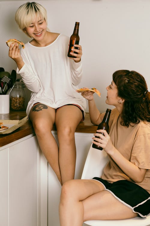 Free Happy female hipster enjoying slice of pizza and drinking beverage while talking to happy friend sitting with crossed legs on chair in apartment Stock Photo