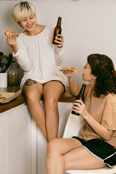 快乐的女潮人一边吃披萨、喝饮料，一边和盘腿坐在公寓椅子上的朋友愉快交谈