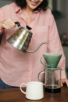 A woman smiles while preparing a fresh pour over coffee in a casual home kitchen setting.
