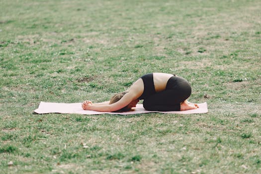 A woman practicing yoga outdoors in a park, in a peaceful child's pose on a mat.