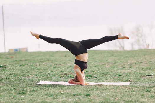 Side view of young sportswoman in black sportswear practicing headstand pose with apart legs on sport mat during outdoors workout adhering to healthy lifestyle