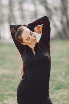 Positive young female in black sportswear standing in park and stretching arms during outdoors training while smiling with closed eyes
