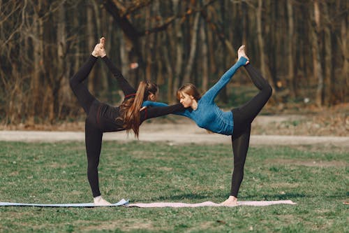 Side view of flexible female friends standing in Natarajasana pose while practicing yoga together and supporting each other on mat in park in daylight