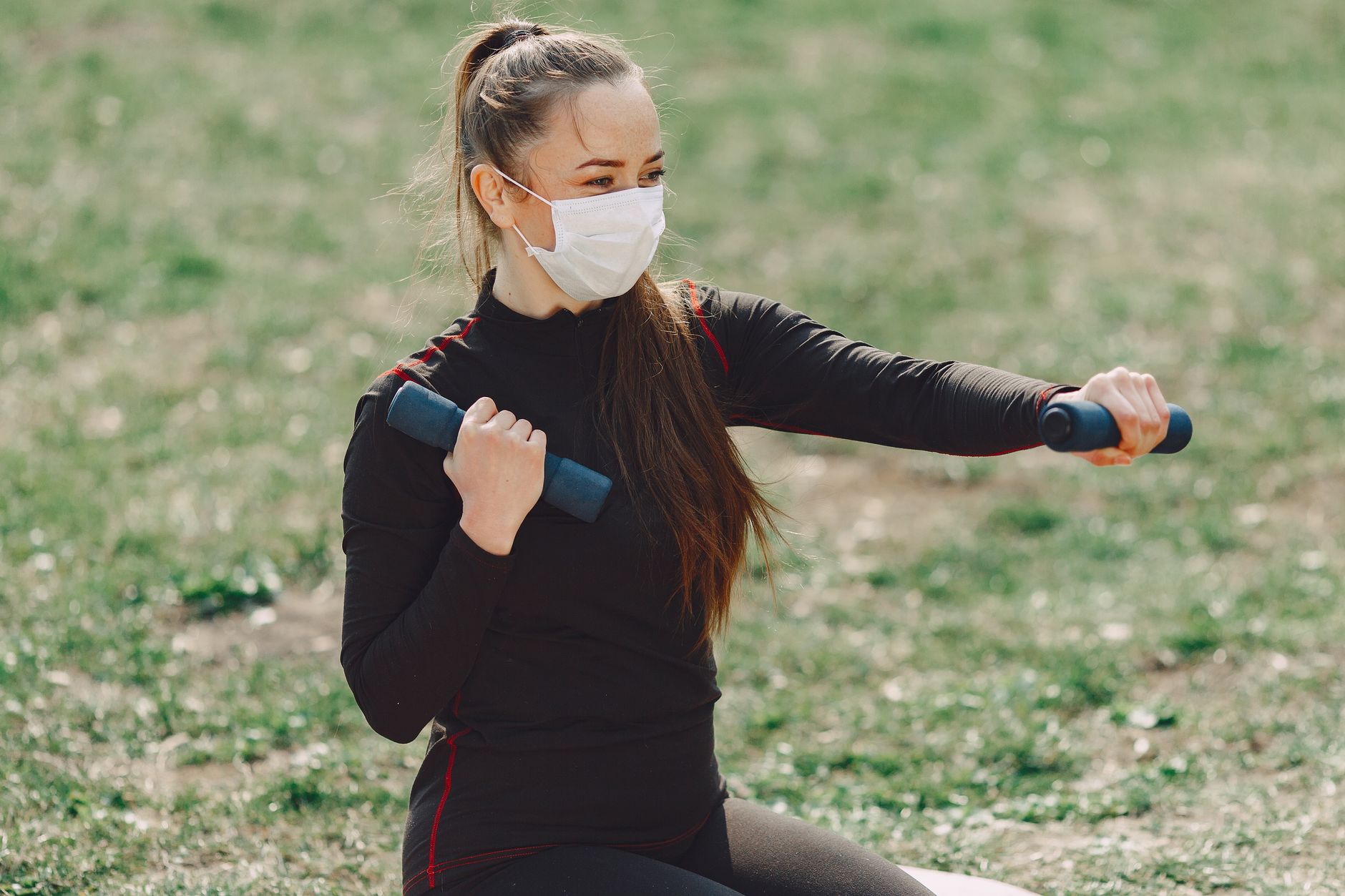 Slim female athlete in sports clothes and sterile mask using dumbbells during workout on grass and looking away in daylight during COVID 19 pandemic