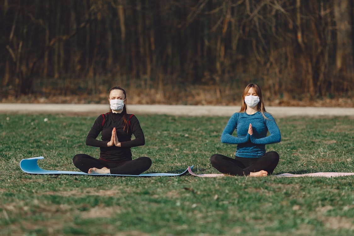 Tranquil girlfriends in face masks sitting in Namaste pose