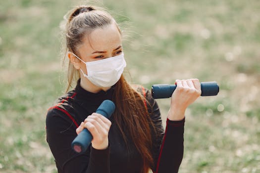 Slender young lady in face mask and sports clothes looking away while exercising with dumbbells outdoors in daylight during quarantine time