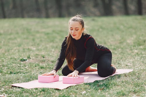 Slender female in sportswear with long hair leaning forward while touching yoga bricks on mat put on grass in park