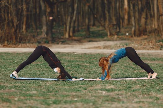 Side view of fit woman in face mask standing in Adho Mukha Svanasana pose near slender unrecognizable female friend on mats put on grass near forest during COVID 19 pandemic