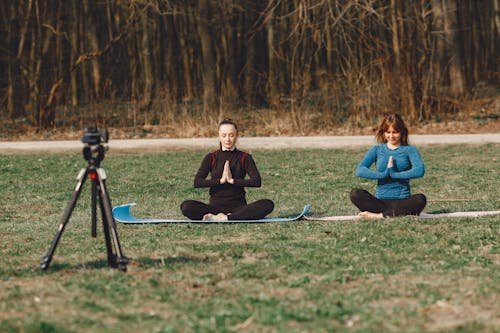 Calm young female friends in sportswear sitting with crossed legs and closed eyes in Namaste pose in front of tripod with photo camera on grass