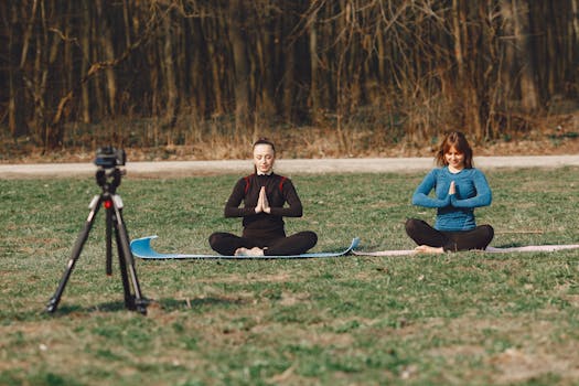 Calm young female friends in sportswear sitting with crossed legs and closed eyes in Namaste pose in front of tripod with photo camera on grass
