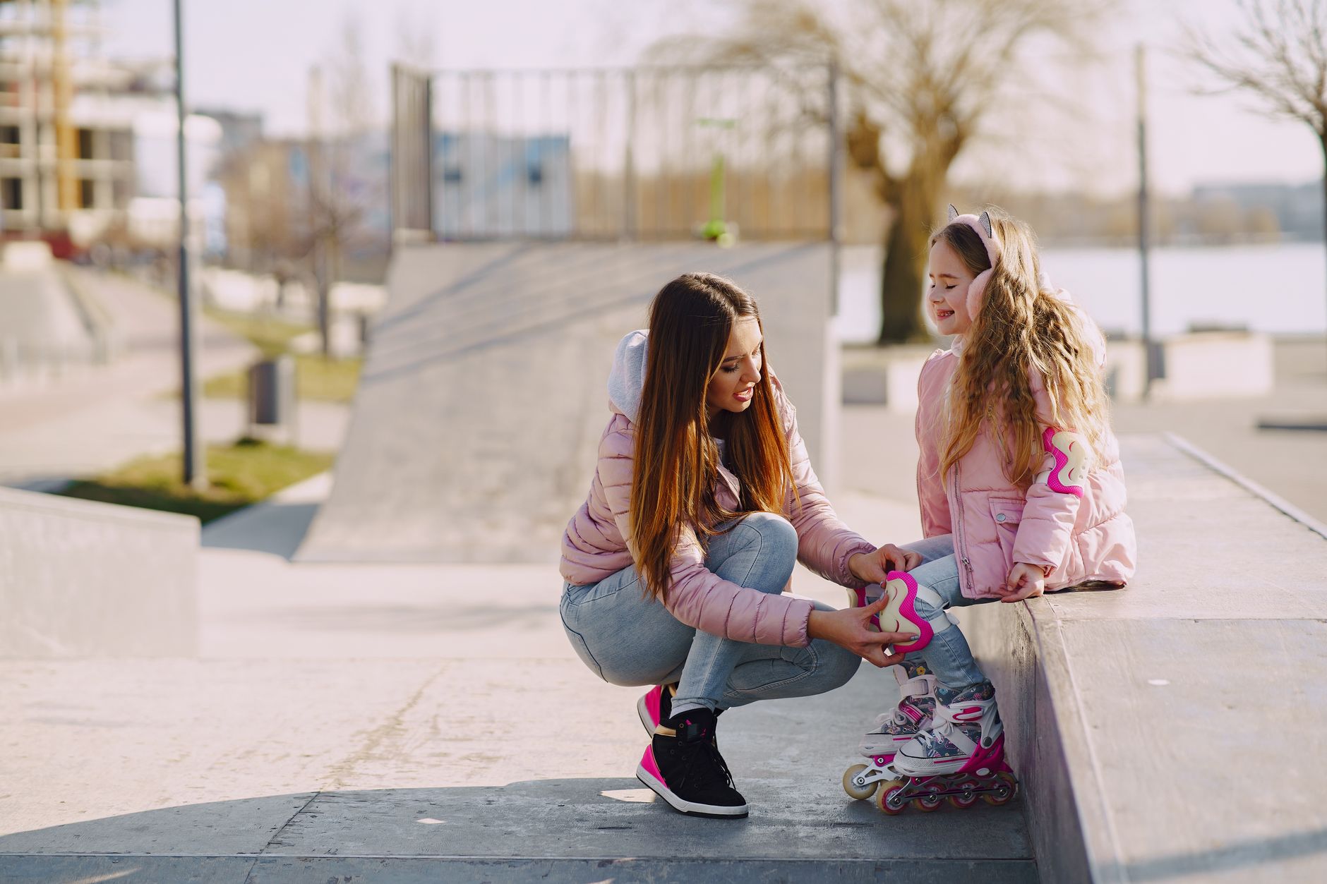 Cheerful young woman adjusting knee pads for little girl during active sport and game outside in city while spending time together