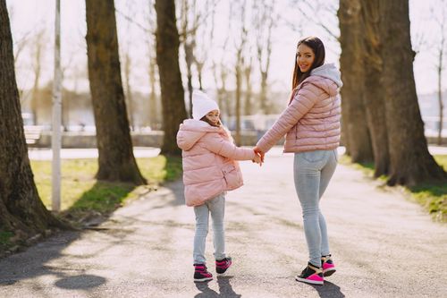 Free Back view of delighted young woman in warm pink jacket smiling and looking at camera over shoulder while walking on path among trees in park in sunny spring day together with little adorable girl Stock Photo