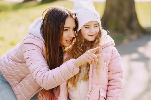 Smiling young woman in warm pink jacket leaning towards adorable girl while pointing at something and looking away with curiosity during stroll in city park in sunny spring day