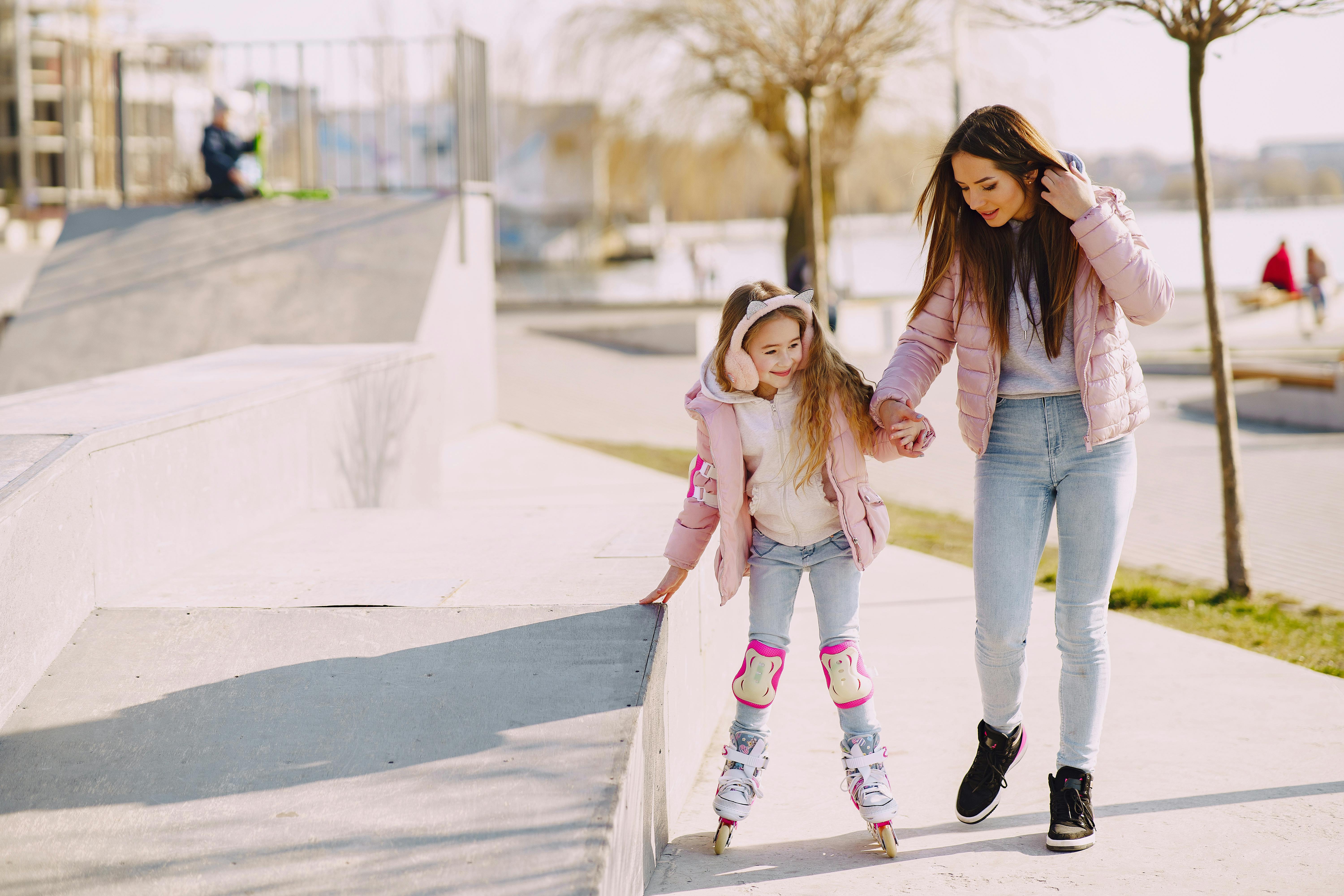 Focused woman in warm clothes supporting little adorable daughter wearing fur earmuffs and protection pads on knee and elbows during roller skating on skate playground with ramps in sunny spring day