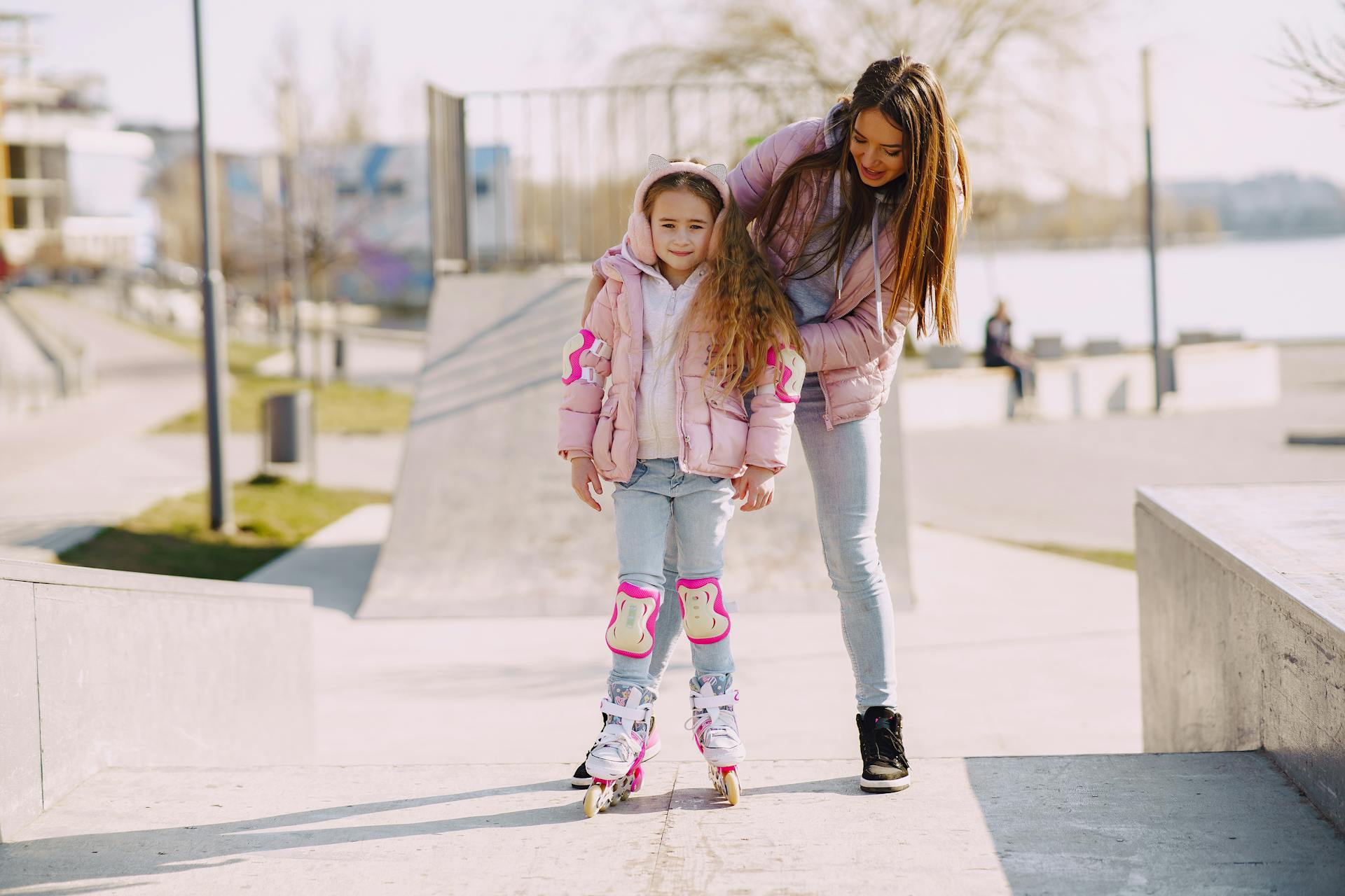 Full body cheerful young woman supporting daughter wearing protection pads while roller skating on concrete ramp during active leisure in sunny spring day