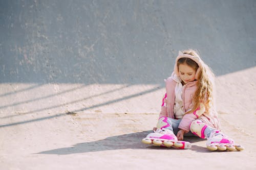 Glad little girl wearing warm pink jacket and protective pads looking down on rollers while sitting on concrete surface of ramp for skating during visiting skate park in sunny cold day