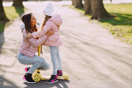 Free Side view of little girl in casual warm wear hugging mother while riding kick scooter on asphalt alley in city park and looking to each other Stock Photo