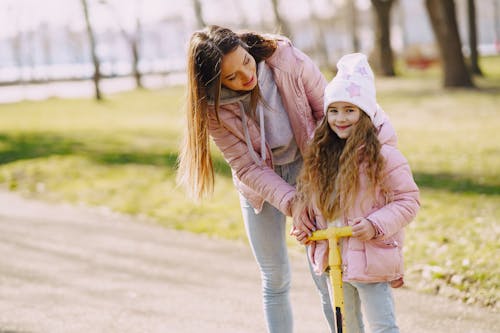 Free Cheerful little girl in warm clothes and hat riding kick scooter with mother help on pathway in park in spring Stock Photo