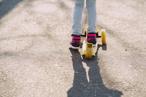Free Back view of crop unrecognizable little child in jeans and sneakers ridding kick scooter on asphalt path in sunny day Stock Photo