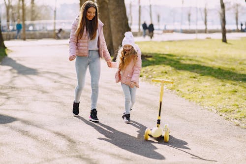 Free Full length female and little girl in warm clothes holding hands while walking and playing on walkway in city park near trees in sunny day Stock Photo