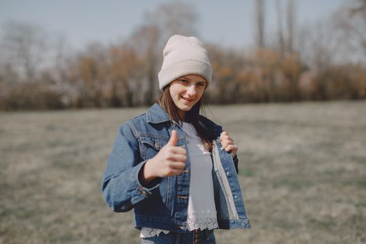 Positive teen girl in jean jacket and hat looking at camera and showing thumbs up at daytime in countryside