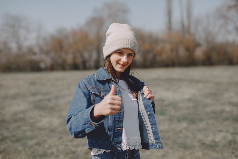 Positive teen girl in jean jacket and hat looking at camera and showing thumbs up at daytime in countryside