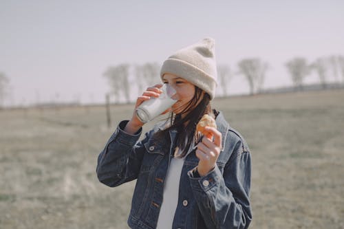 Free Satisfied teen girl in jean jacket and cap drinking milk and holding cookies in hand while spending time in nature Stock Photo