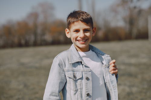 Free Delighted modern kid in jeans jacket standing on field and smiling at camera while having trip in countryside in spring Stock Photo