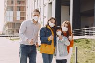 Optimistic family in protective masks and casual clothes standing together on street and showing thumbs up gesture while looking at camera