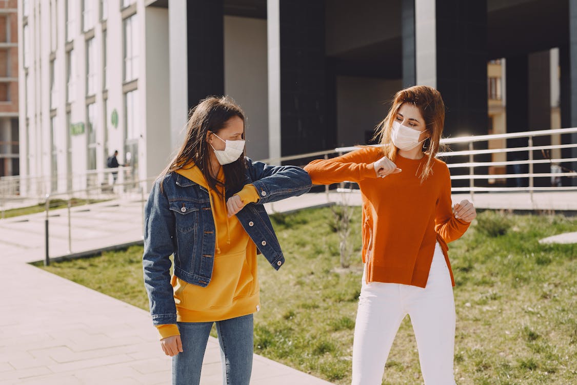 Young women in medical masks greeting each other on city street