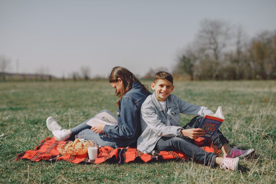 Smiling young boy in casual clothes sitting together with girl and reading interesting story while leaning back to back on checkered blanket on grass in park